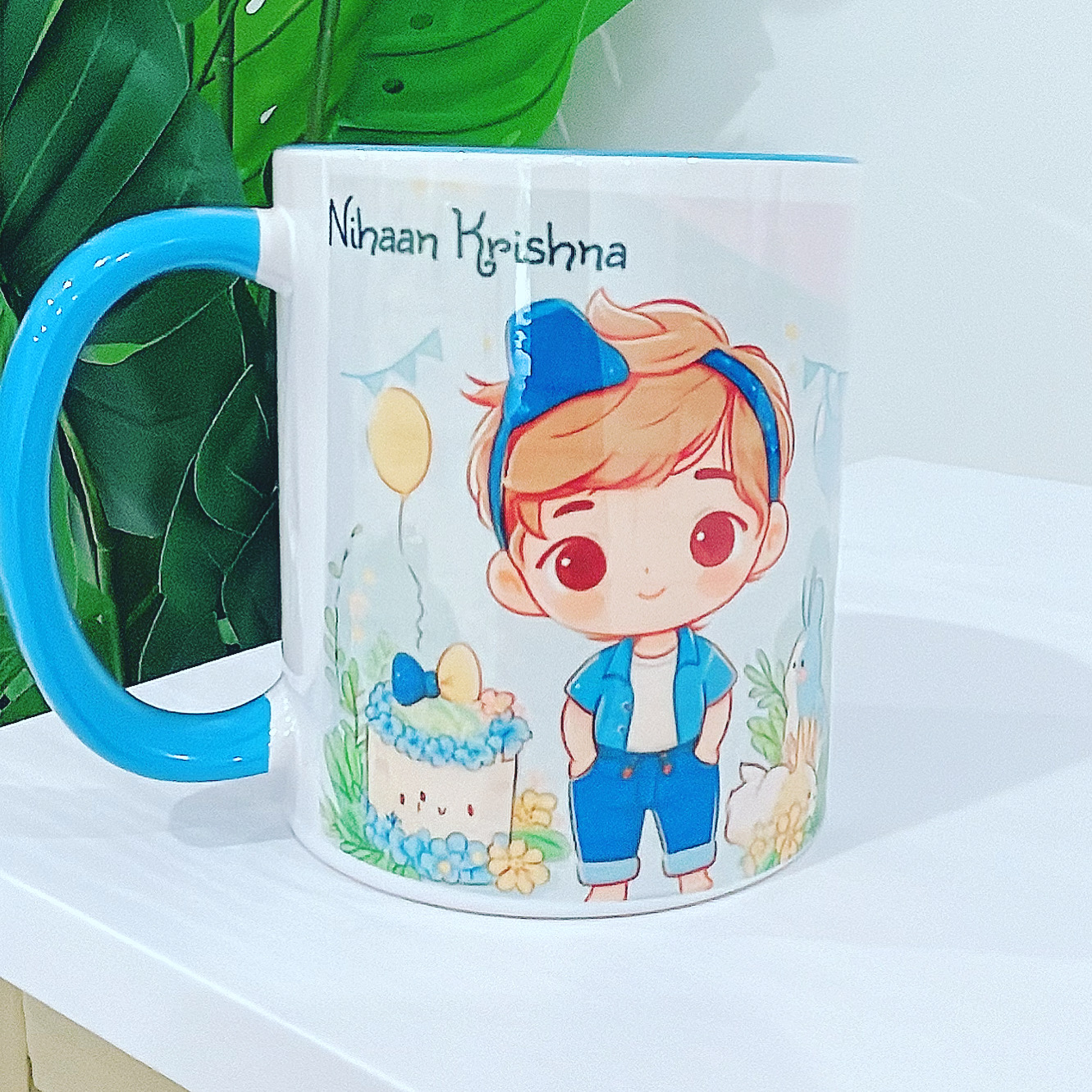 Personalized birthday mug with cute cartoon boy illustration, balloons, cake, and the name "Nihaan Krishna" on a white and blue ceramic cup.
